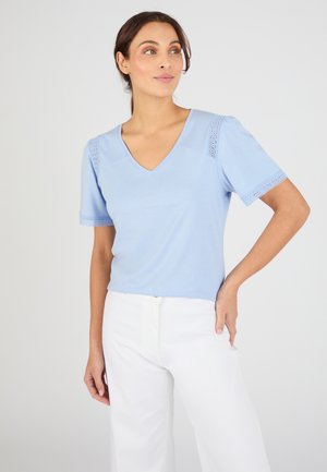 Donna con capelli castani, indossa un top azzurro chiaro a scollo a V con dettagli in pizzo sulle maniche e pantaloni bianchi, in piedi con una mano sul fianco.