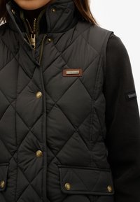 Zwarte gewatteerde vest met hoge kraag, voorzien van gouden knopen, een bruine leren logopatch en zwarte fleece mouwen. Verstelbare ritssluiting.