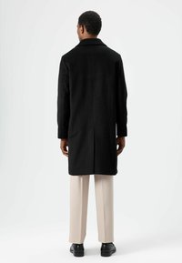 Zwarte jas met een getailleerd ontwerp, lange mouwen en een rechte snit. Draagt over lichtbeige broek en zwarte schoenen. Eenvoudige, gestructureerde stof.
