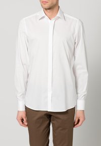 Mann trägt ein tailliertes weißes Hemd mit langen Ärmeln und verdeckten Knöpfen sowie eine braune Hose, steht vor einem schlichten Hintergrund.