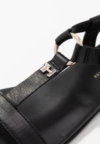 Svart läder sandaler med platt sula, ankelrem med en metallring och en silverfärgad logotypaccent på remmen.