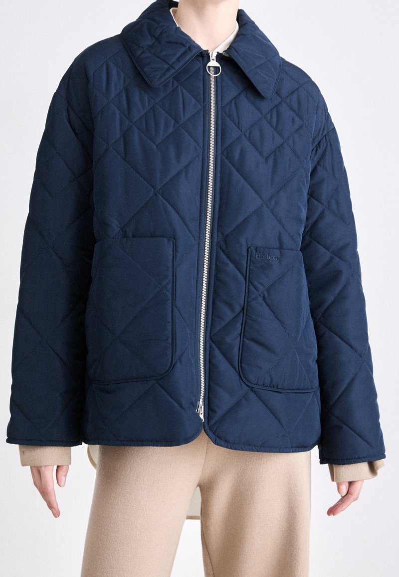 Personne portant une veste matelassée bleu marine à fermeture éclair avec grand col et poches plaquées avant, sur un pantalon beige.