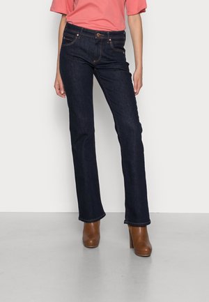 Woman wearing dark blue straight-leg jeans, brown high-heeled ankle boots, and a coral short-sleeve top, standing on gray floor.