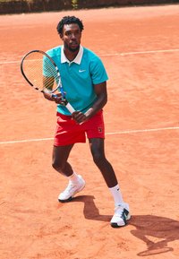 Tennis player in a turquoise polo and red shorts, holding a racket. White athletic shoes with black accents, playing on a clay court.