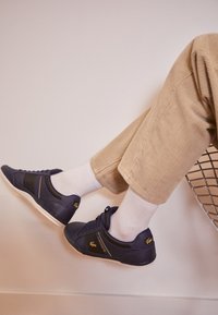 Granatowe skórzane sneakersy z czarnymi teksturowanymi akcentami, białymi podeszwami i złotym logo "LACOSTE". Połączone z beżowymi sztruksowymi spodniami i białymi skarpetkami.