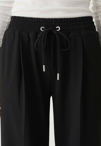 Zwarte elastische broek met een trekkoordsluiting, voorzien van zilverkleurige metalen punten en een gladde, gestructureerde stofafwerking.