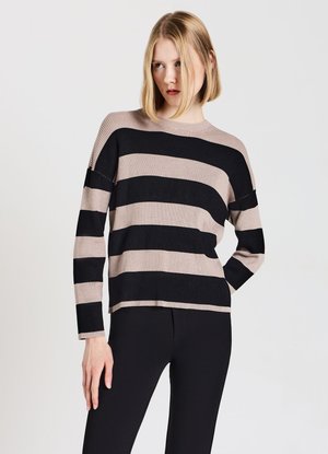 Frau mit kurzen blonden Haaren, die einen lockeren, beige-schwarz breit gestreiften Pullover und schwarze Hose vor einem schlichten hellen Hintergrund trägt.