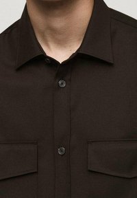 Chemise marron à boutons avec col, équipée de deux poches poitrine et de boutons noirs, confectionnée dans un tissu tissé lisse.