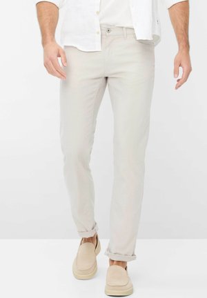 Mann trägt beige schmal geschnittene Hose mit hochgekrempelten Säumen, passende beige Slipper und ein weißes Hemd mit hochgekrempelten Ärmeln.