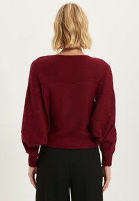Pull en tricot bordeaux à coupe décontractée, manches bouffantes, et motif texturé. Dispose d'un encolure large et d'un dos ouvert.