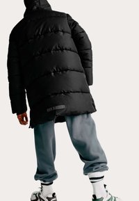 Zwarte pufferjas met doorgestikte panelen en een logo patch, gecombineerd met grijze sweatpants en gemêleerde sokken, getoond vanuit de achterkant.