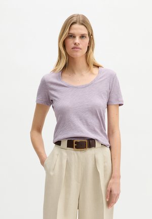 Blonde Frau, die ein helllila kurzärmeliges Hemd trägt, das in eine hoch taillierte beige Hose mit einem dunkelbraunen Gürtel gesteckt ist.