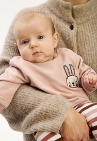 Bluza pentru bebeluși roz deschis cu grafică de iepuraș, decolteu rotund și capse. Colanți maro cu dungi. Pulover gri texturat în fundal.