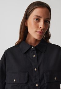 Camicia nera con bottoni, realizzata in tessuto testurizzato, caratterizzata da un colletto classico, due tasche sul petto e bottoni a pressione metallici.