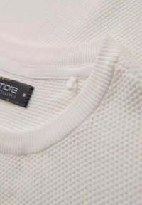 Tejido de punto blanco texturizado con cuello acanalado y pequeño detalle de lazo. La etiqueta adjunta muestra el nombre de la marca y la talla.