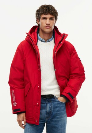 Mand iført en lys rød vinterjakke over en hvid sweater og blå jeans med den ene hånd i lommen mod en ensfarvet baggrund.