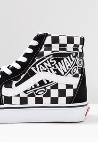 Vysoké tenisky Vans s černobílým šachovnicovým vzorem, bílé tkaničky, logo a bílá gumová podrážka.