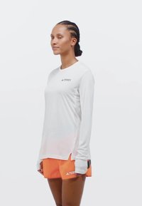 Camisa atlética de manga larga blanca con textura de malla, combinada con pantalones cortos naranja brillante que cuentan con bolsillos laterales y el logo de la marca.
