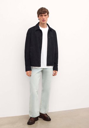Chaqueta de ante negra sobre una camiseta blanca, combinada con jeans de color azul claro y zapatos gruesos marrones oscuros, de pie contra un fondo neutral.