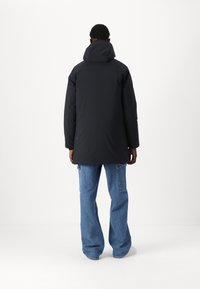 Giubbotto nero isolato con cappuccio, caratterizzato da una silhouette dritta e maniche lunghe. Indossato sopra jeans a gamba larga blu chiaro.
