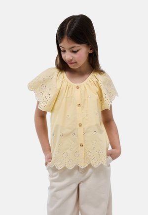 Fille aux longs cheveux foncés porte un chemisier jaune à boutons œillets avec des bordures festonnées et un pantalon clair, les mains dans les poches, regardant vers le bas.