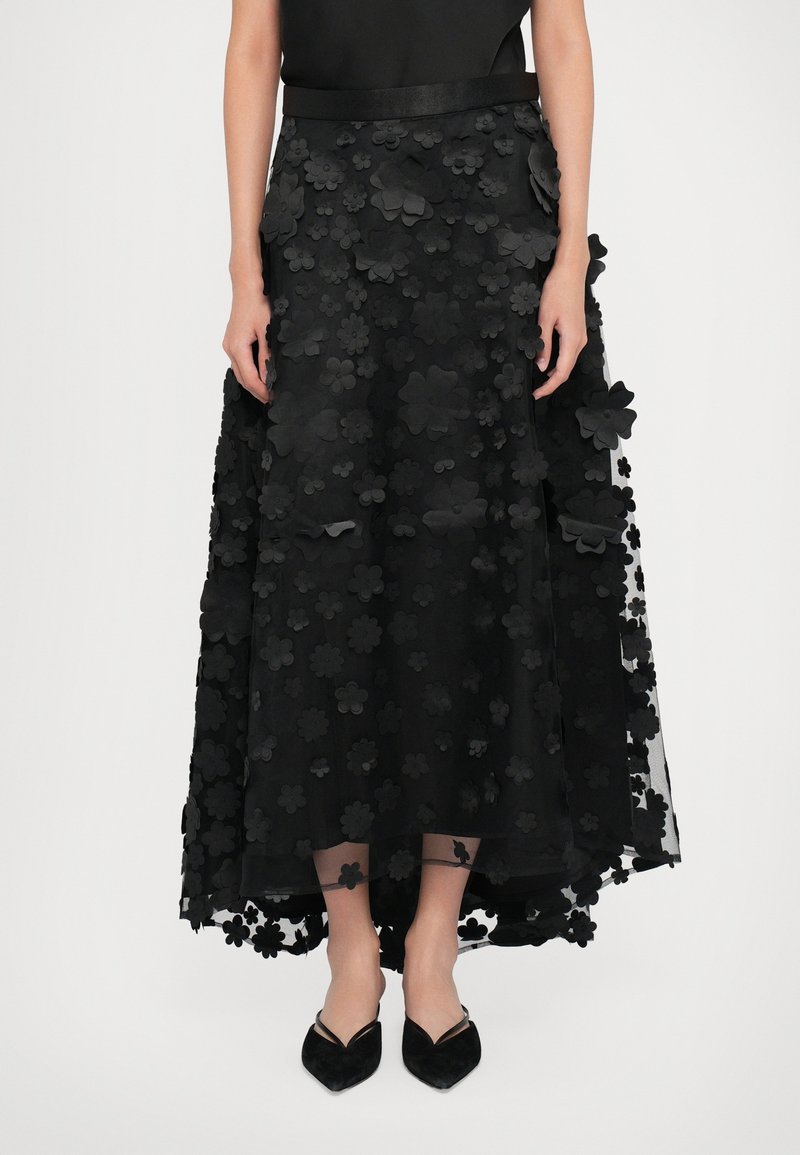 Jupe longue noire avec des appliques florales 3D superposées, portée avec des chaussures noires pointues et un détail floral transparent près des chevilles.