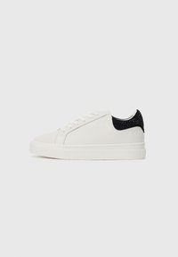 Hvite lave sneakers med snøring foran og svart hælflik pyntet med svarte nagler, satt mot en enkel hvit bakgrunn.