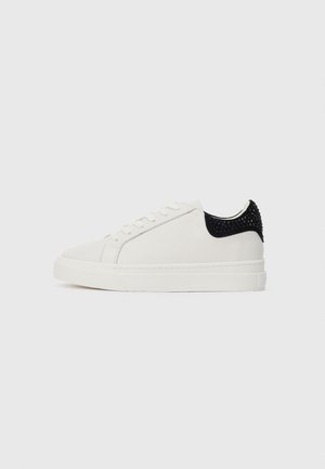Sneaker bassa bianca con allacciatura frontale e tallone nero decorato con borchie nere, posizionata su uno sfondo bianco semplice.