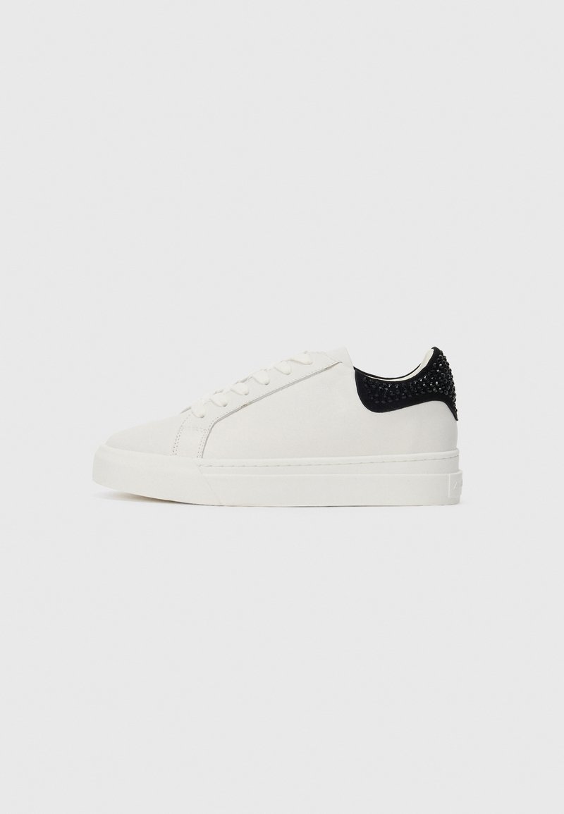 Hvite lave sneakers med snøring foran og svart hælflik pyntet med svarte nagler, satt mot en enkel hvit bakgrunn.