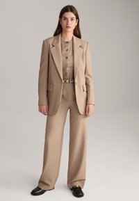 Beiger maßgeschneiderter Blazer über einem braunen Hemd-ähnlichen Pullover, kombiniert mit weiten beigen Hosen und schwarzen Schuhen. Enthält einen dekorativen Gürtel.