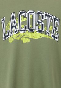 Olīvu zaļš kokvilnas T-krekls ar izteiksmīgu "LACOSTE" tekstu tumši zili un stilizētu dzeltenu krokodilu grafiku zem tā.