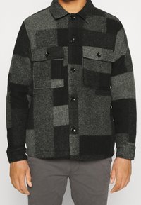 Hombre vistiendo una chaqueta de lana abotonada a cuadros en negro y gris con dos bolsillos en el pecho y pantalones grises.