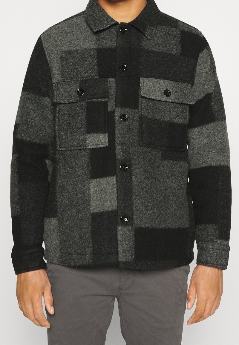 Hombre vistiendo una chaqueta de lana abotonada a cuadros en negro y gris con dos bolsillos en el pecho y pantalones grises.