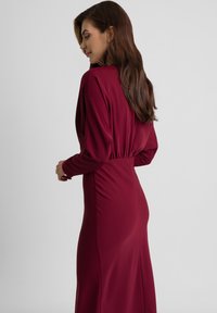 Burgundowa sukienka z długim rękawem, wykonana z miękkiego materiału, z wysmuklającym paskiem w talii i gładkim, płynącym krojem o lekkiej teksturze.