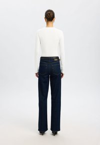 Tummansiniset farkut, valmistettu denimistä, suora lahje, jossa on takataskut ja musta vyö. Pitkähihainen valkoinen paita näkyy selästä.