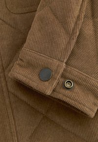 Manche de veste en velours côtelé marron avec des boutons-pression texturés visibles, un fermé et un ouvert, montrant des plis détaillés du tissu et les coutures.