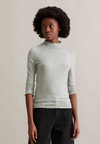 Slim-fit Langarmoberteil aus hellgrünem Rippstoff, mit einem hohen Kragen mit gewelltem Saum und umgeschlagenen Ärmeln. Kombiniert mit schwarzen Hosen.