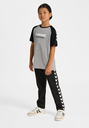 Grå T-shirt med sorte raglanærmer og logo tekst, kombineret med sorte bukser med hvide chevronstriber ned langs siderne og hvide sneakers.