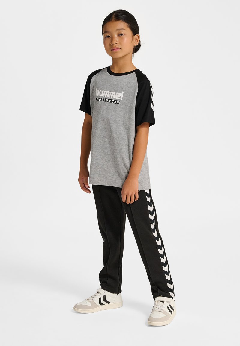 Szara koszulka z czarnymi rękawami typu raglan i tekstem z logo, zestawiona z czarnymi spodniami z białymi paskami w kształcie litery V po bokach oraz białymi trampkami.