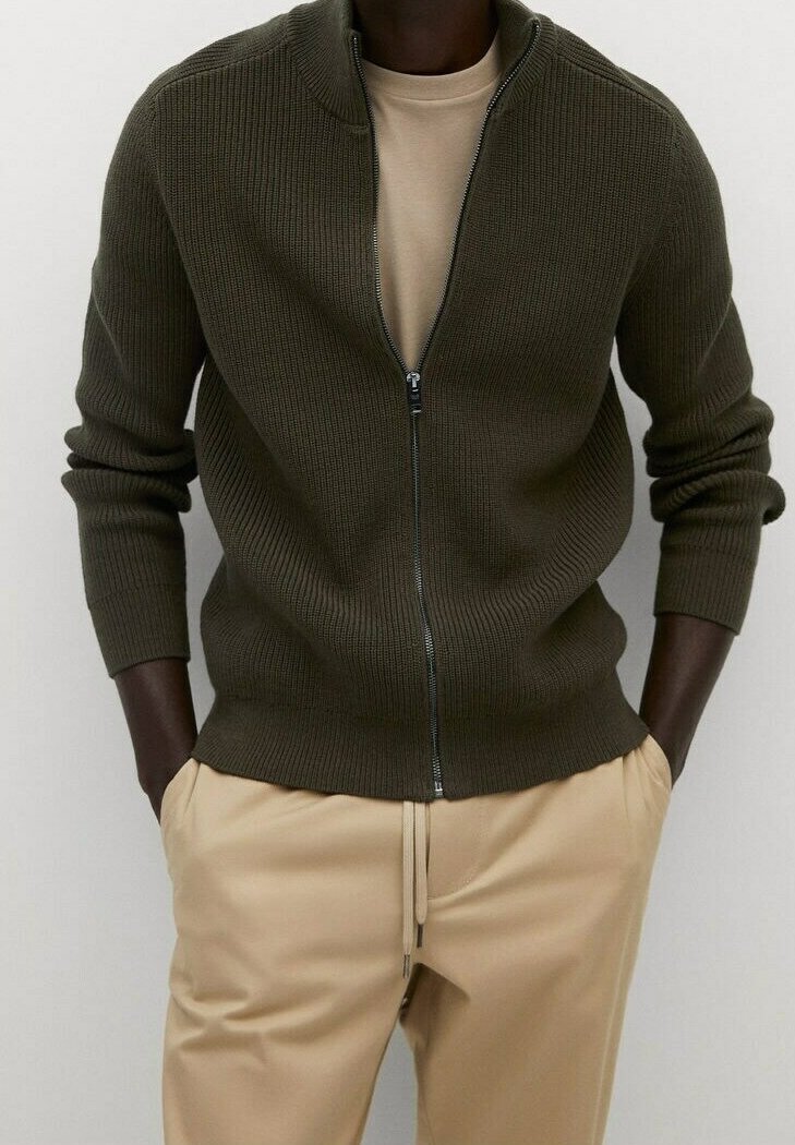Cárdigan con cremallera en tejido de punto acanalado verde oscuro, con cuello alto y mangas largas, combinado con pantalones beiges.