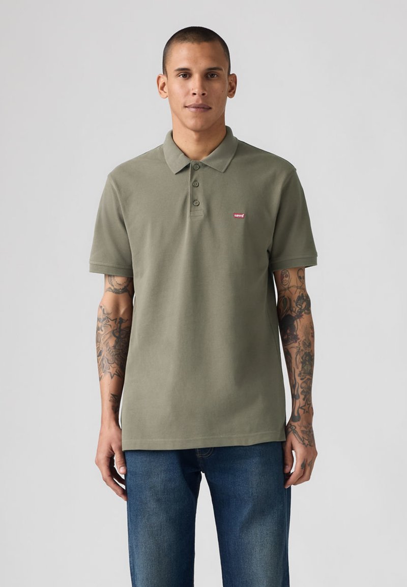 Homme tatoué portant un polo Levi's vert olive et un jean bleu, debout devant un fond gris uni.