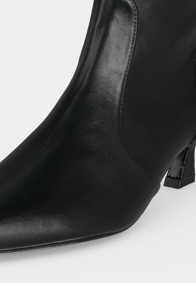 Bottine noire en cuir avec un bout pointu et un petit talon bloc sur un fond clair uni.