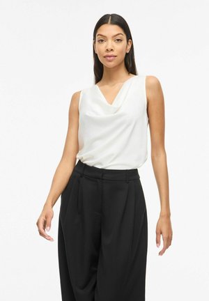 Donna con capelli lunghi e scuri che indossa una camicetta bianca senza maniche e pantaloni neri a gamba larga e vita alta, in piedi davanti a uno sfondo semplice.