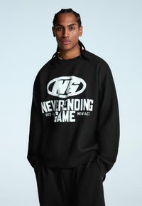 Čierny oversized sveter vyrobený z mäkkej tkaniny, ktorý má bielu grafiku s textom "NEKONČIACE HRY" a logom.