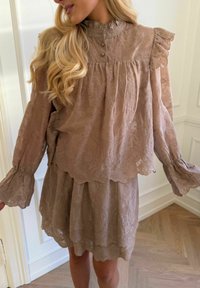 Žena u svijetlosmeđoj haljini od čipke s naboranim detaljima na ramenima i dugim proširenim rukavima, stoji unutra na drvenom podu.