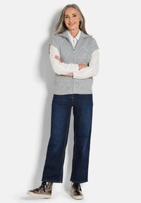 Gilet sans manches en tricot gris avec une fermeture éclair, porté sur une chemise blanche à col, associé à un jean large bleu foncé et des baskets métalliques.