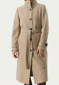 Cappotto in misto lana beige con colletto alto, sei bottoni neri e una cintura abbinata in vita. Presenta maniche lunghe con polsini piegati.