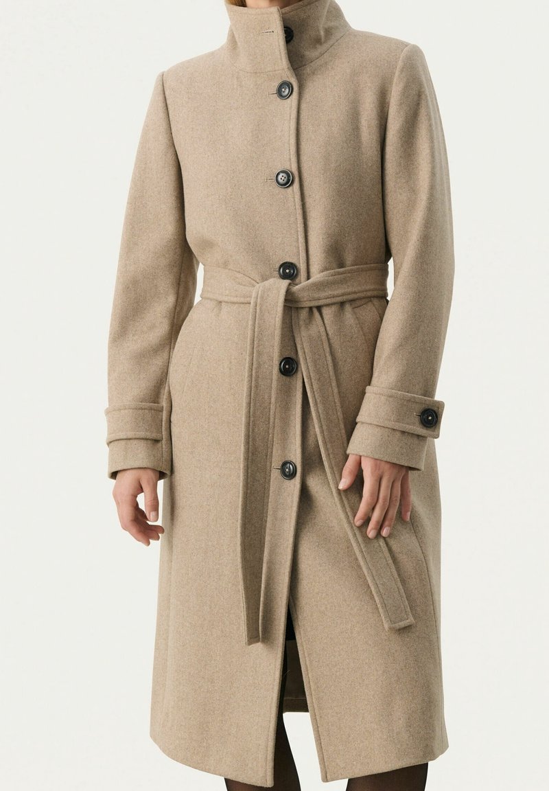 Cappotto in misto lana beige con colletto alto, sei bottoni neri e una cintura abbinata in vita. Presenta maniche lunghe con polsini piegati.
