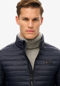 Granatowa kurtka puchowa z zamkiem z przodu, textured fabric, i stójką, noszona na jasnoszarym, prążkowanym golfie. Logo na lewej piersi.