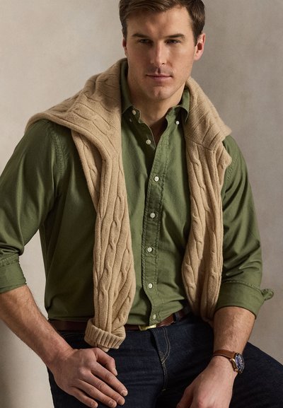 Mladý muž oblečený v olivovo zelenej košeli na gombíky s béžovým vrkočovým svetríkom prehodeným cez ramená, tmavé džínsy a hodinky na zápästí, sediaci s rukami položenými.